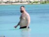 Sam Smith sem camisa mostra seu torso tatuado enquanto um amigo passa protetor photo voltaic em suas costas durante o dia à beira-mar após um present no México